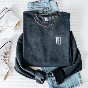 Peut inclure: Un sweat-shirt gris foncé avec un chiffre romain VII brodé en blanc sur le devant. Le sweat-shirt est posé sur une surface blanche avec une paire de baskets blanches et un jean bleu.