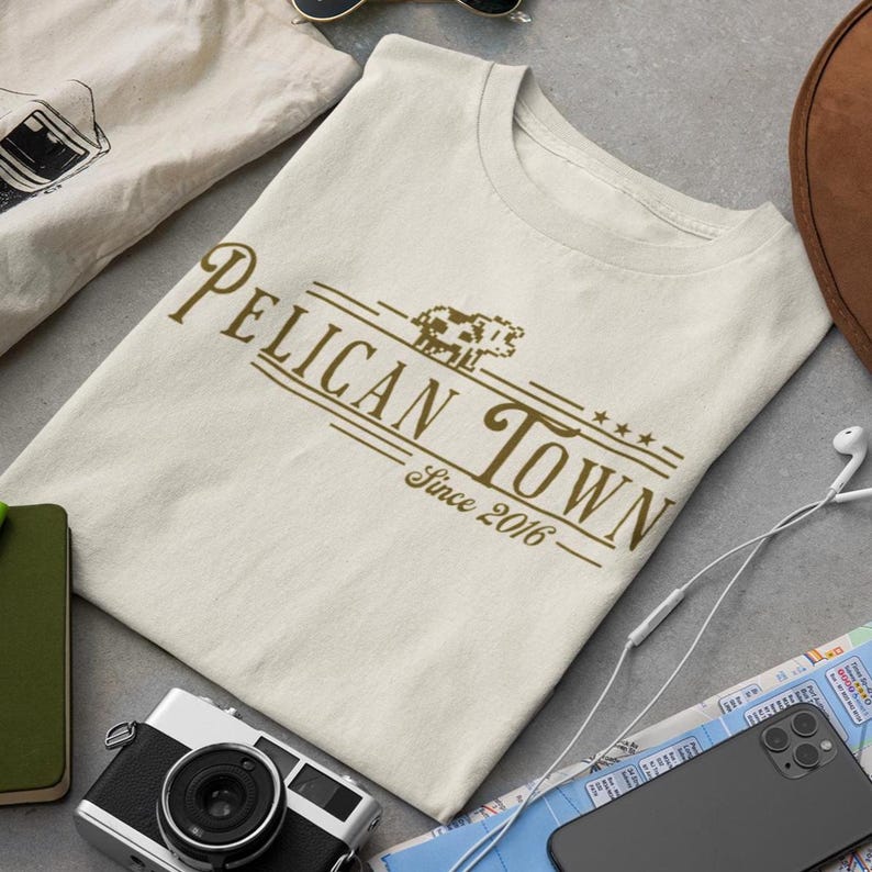 Op de afbeelding: Cr&egrave;mekleurig T-shirt met de tekst "Pelican Town Since 2016" in bruin. Het ontwerp bevat een gestileerde koeiengrafiek. Het shirt wordt gepresenteerd met reisaccessoires.