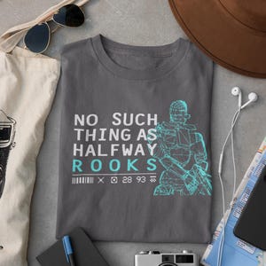 Puede incluir: Camiseta gris con el texto "NO SUCH THING AS HALFWAY ROOKS" y un gráfico turquesa de una figura futurista. Accesorios: gafas de sol, sombrero, cámara y cuaderno.