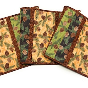 May include: A set of four rectangular quilted placemats featuring a repeating pattern of brown pinecones and green pine needles. The fabric panels alternate between light tan and dark green backgrounds, all bordered by a solid brown fabric trim.