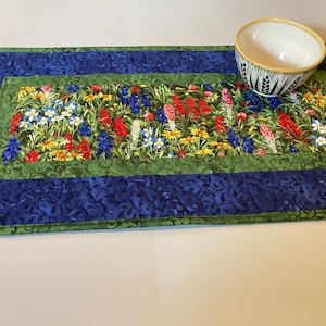 May include: A rectangular quilted table runner with a floral design. The runner features a central panel with colorful wildflowers, framed by green and blue patterned borders. A white and black bowl sits on the runner.