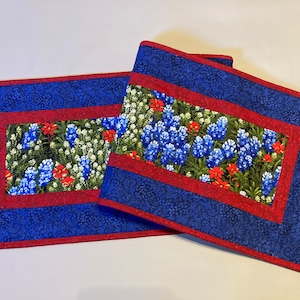 May include: Two rectangular placemats with a floral design. The center features a field of bluebonnets, red flowers, and white flowers. The border is a deep blue with a swirling pattern, framed by a red edge.