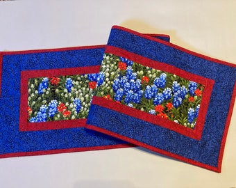 May include: Two rectangular placemats with a floral design. The center features a field of bluebonnets, red flowers, and white flowers. The border is a deep blue with a swirling pattern, framed by a red edge.