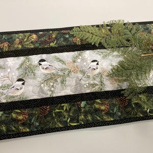 May include: A decorative table runner featuring a winter scene with chickadee birds, pine branches, and pine cones. The runner has a black border with a dotted pattern and a green and brown pine cone design.