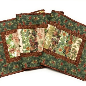 May include: Set of three quilted placemats. Each placemat features a central panel with pine cones and evergreen branches on a cream background, framed by a brown border and a green outer border with pine cone accents. A rustic, seasonal theme.