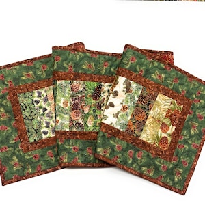 May include: A set of three rectangular fabric placemats featuring a patchwork design with a green background, brown borders, and a repeating pattern of pinecones, pine needles, and evergreen branches in shades of brown, green, and cream.