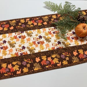 May include: A fall-themed table runner with a leaf pattern in shades of orange, yellow, and brown. The center panel features a white background with scattered leaves. A small pumpkin and greenery are placed on top.