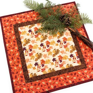 May include: A square quilted fabric mat featuring a central panel of cream fabric with a colorful autumn leaf print, surrounded by a dark brown border and an outer border of orange fabric patterned with small acorns and leaves.