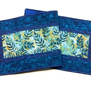 May include: Two rectangular quilted placemats. The placemats have a dark blue plaid border and a central panel with a leaf pattern in shades of green and yellow. The placemats are made of fabric and are likely handmade.