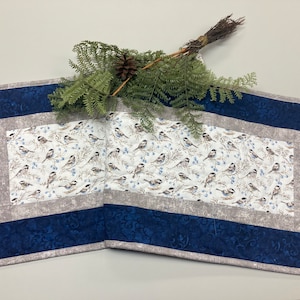 May include: A blue and gray quilted table runner with a white background featuring a pattern of small birds. The runner has a decorative border with a blue and gray pattern.