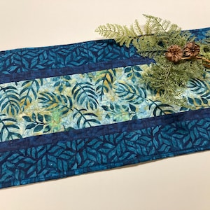 May include: A rectangular table runner with a leaf pattern in shades of blue and yellow. The runner has a dark blue border with a leaf design. A sprig of greenery and pinecones rests on the runner.