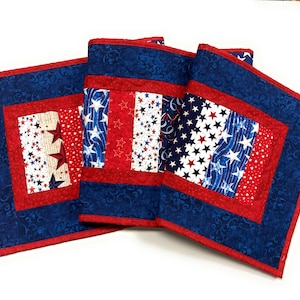 May include: A set of patriotic quilted placemats in red, white, and blue. The placemats feature a square design with a red border and a central panel with star patterns. The blue fabric has a swirling pattern.