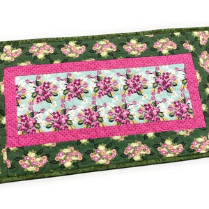 May include: A rectangular quilted table runner with a floral design. The runner features a central panel with a light blue background and pink flowers. It is framed by a pink border and a green floral patterned border. The edges are finished with a green binding.