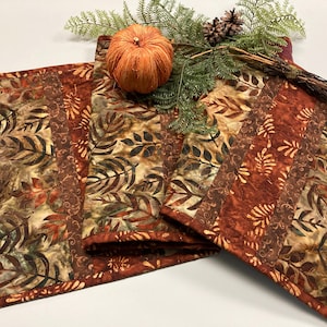 May include: A fall-themed table runner with a leaf pattern in shades of brown, orange, and green. An orange pumpkin, pinecones, and greenery are arranged on top. The runner is made of fabric and is likely used for home decor.