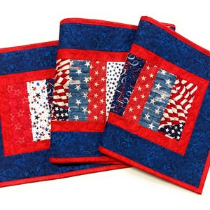 May include: A quilted table runner and two matching placemats featuring a patriotic theme. The fabric displays red, white, and blue colors with patterns of stars, stripes, and swirling designs. Each piece is bordered by a solid red fabric frame.