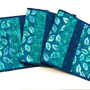 May include: A set of three rectangular quilted placemats featuring a patchwork design in shades of teal, turquoise, and navy blue. The fabric patterns include stylized leaves and starburst motifs with dark blue borders and intricate stitching.