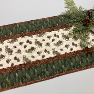May include: A quilted table runner with a forest theme. The runner features a central cream-colored panel with pinecone and branch patterns, bordered by bands of green trees and brown patterned fabric. Ferns and pinecones are placed on top.