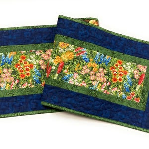 May include: Two rectangular quilted placemats. The center features a vibrant floral pattern with red, yellow, blue, and pink flowers on a green background. A dark blue border frames the floral design, with a green border in between.