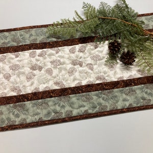 May include: A brown and green table runner with a pine cone pattern. The runner has a brown and green border with a decorative pattern. The runner is on a white surface with a sprig of greenery and pine cones.