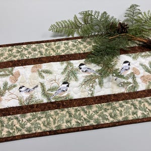 May include: A rectangular quilted table runner featuring a pattern of black-capped chickadees, pine branches, and pinecones on a cream background, bordered by dark brown fabric strips and a green pine needle print fabric along the edges.