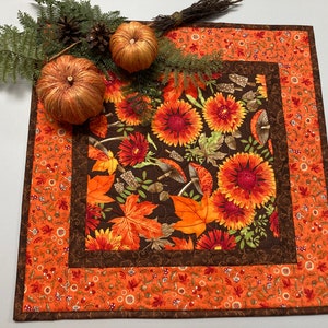 May include: A square quilted table runner with a fall theme. The center features a dark brown fabric with orange and red flowers and leaves. The border is orange with a floral pattern. Two small pumpkins and greenery are on top.