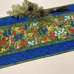 May include: A green and blue table runner with a floral pattern featuring bluebonnets, red Indian paintbrush, and other wildflowers. The runner has a green border with a blue and green patterned border.