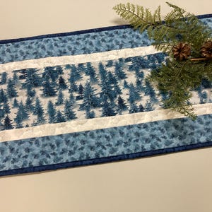 May include: A rectangular table runner with a winter forest design. The center features a white background with blue trees. The edges are blue with a floral pattern, and a dark blue border. A sprig of greenery and pinecones are on the right.