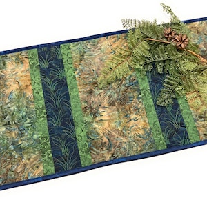 May include: A rectangular table runner with a blue border and a quilted design. The runner features vertical stripes in shades of green, blue, and brown. A decorative sprig of greenery with pinecones rests on the runner.