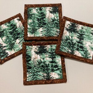 May include: Set of four square fabric coasters. Each coaster features a forest scene with green and white trees, framed by a brown border with a decorative pattern. The coasters are designed to protect surfaces from heat and moisture.
