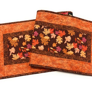 May include: Two rectangular quilted table runners featuring a central panel with a dark brown background printed with orange, red, and yellow autumn leaves, bordered by a textured orange fabric with a subtle swirling leaf pattern.