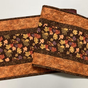 May include: Two quilted placemats with a fall theme. The placemats feature a brown border with an orange floral pattern. The center of the placemats have a dark brown background with colorful leaves in shades of orange, yellow, and purple.