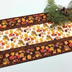 May include: A quilted table runner featuring a central panel with a pattern of autumn leaves in shades of orange, yellow, red, and brown on a cream background, bordered by dark brown fabric strips decorated with a matching leaf print design.