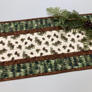 Puede incluir: Camino de mesa acolchado con temática de la naturaleza. El corredor presenta un panel central crema con un patrón de piñas marrones y agujas de pino verdes. Los bordes tienen un borde marrón y un patrón de bosque verde.