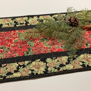 May include: A Christmas-themed table runner with a poinsettia and pinecone design. The runner features a red center with large poinsettias, bordered by black sections with a pinecone pattern. A sprig of greenery and pinecones adorn the top.