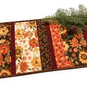 May include: A rectangular quilted table runner featuring a patchwork design of autumn-themed fabrics. The pattern includes sunflowers, orange and red flowers, brown foliage, and acorns on cream, dark brown, and burgundy backgrounds with a pine sprig.