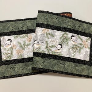 May include: Two quilted table runners. The runners feature a winter scene with chickadees, pine branches, and pinecones on a gray background. The borders are green with a black dotted trim. The runners are approximately 12 inches wide and 24 inches long.