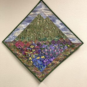 May include: A quilted wall hanging with a diamond shape. The artwork depicts a landscape with a forest, mountains, and a vibrant field of flowers in shades of pink, purple, yellow, and blue. The quilt is framed with a green border.