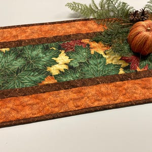 May include: A fall-themed table runner with a central panel of green maple leaves and accents of orange and brown. A decorative pumpkin, pinecone, and greenery complete the autumn display.