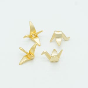 May include: Four small, gold-colored metal beads shaped like stylized origami cranes. Each bead features a smooth, matte finish with pointed wings and a tail, designed with a central opening for threading, presented in various orientations.