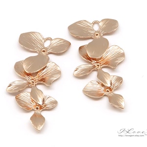 May include: Two gold-tone metal flower charms with a textured finish. Each charm features three overlapping flower shapes with a hole in the center for attaching to jewelry.