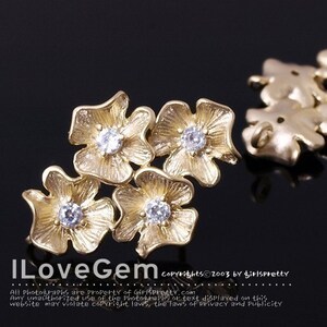 May include: A gold-toned metal connector featuring four flower-shaped components arranged in a cluster, each with a central clear round gemstone. The text ILoveGem and copyright information are visible at the bottom of the image.