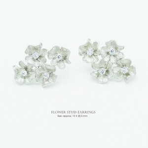 May include: A pair of silver-toned flower stud earrings featuring textured petals and a central clear stone in each flower. The text reads FLOWER STUD EARNINGS Size: approx. 15 X 20.5 mm. These earrings consist of multiple connected floral shapes.