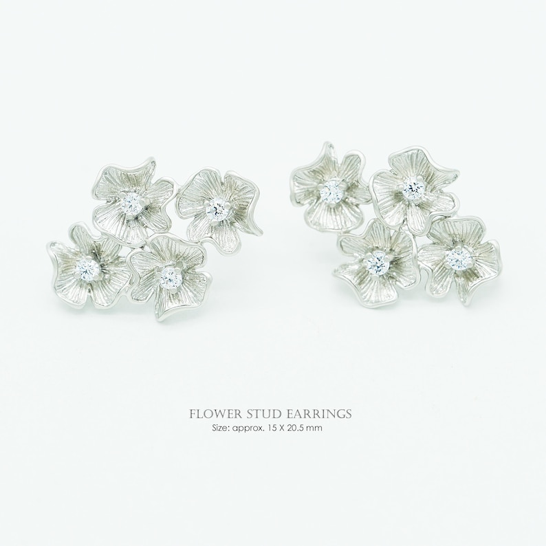 May include: A pair of silver-toned flower stud earrings featuring textured petals and a central clear stone in each flower. The text reads FLOWER STUD EARNINGS Size: approx. 15 X 20.5 mm. These earrings consist of multiple connected floral shapes.