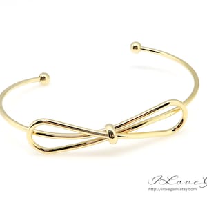 May include: A gold-toned bangle bracelet with a double bow design. The bracelet is open and has a simple, elegant style.
