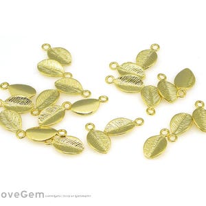 Puede incluir: Encantos de hojas doradas con bucles para hacer joyas. Los encantos tienen patrones detallados de venas de hojas y superficies lisas. Los encantos están dispersos sobre un fondo blanco.