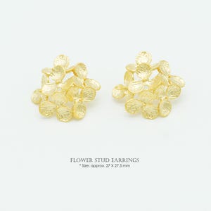 Può includere: Un paio di orecchini a lobo a forma di fiore color oro con un design a petali organici e testurizzati. Gli orecchini sono etichettati come FLOWER STUD EARRINGS con una dimensione di circa 27 x 27,5 mm. La superficie ha una finitura opaca martellata.