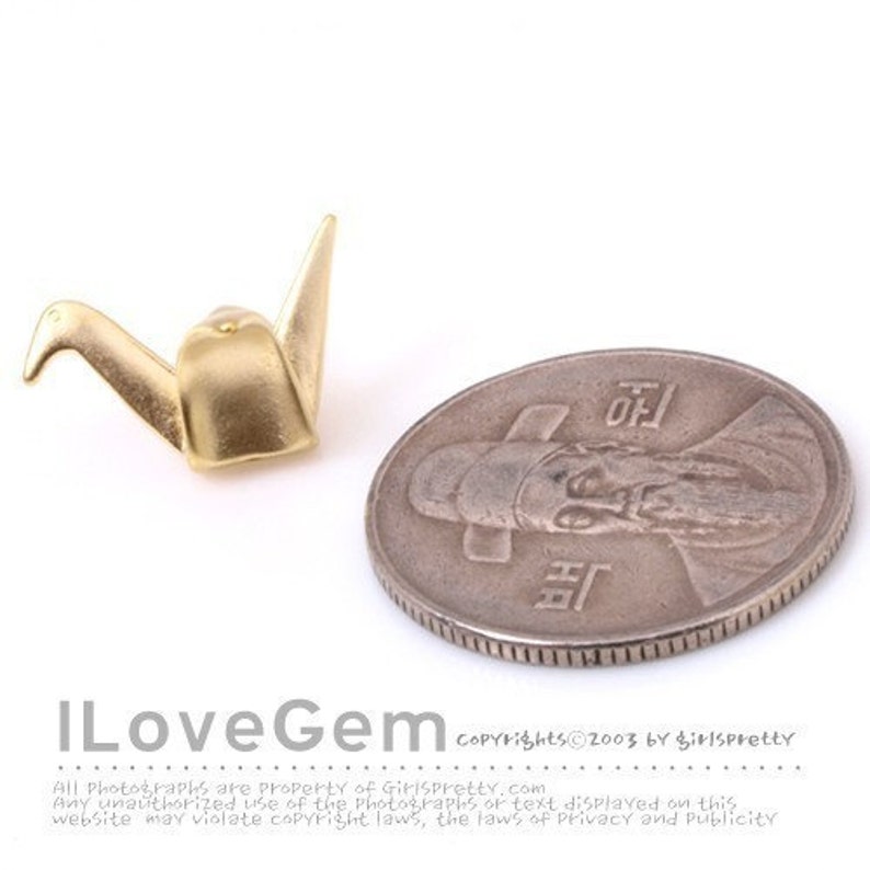 May include: A small gold-colored metal origami crane figurine positioned next to a circular silver-toned coin. The coin features a portrait of a person and Korean text. The text ILoveGem and copyright information are printed at the bottom of the image.