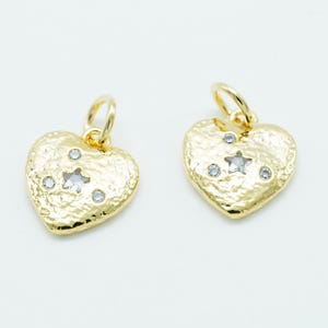 May include: Two gold-colored heart-shaped charms with textured surfaces. Each charm features a star-shaped and two round clear crystal accents. The charms have a loop at the top for attaching to a necklace or bracelet.