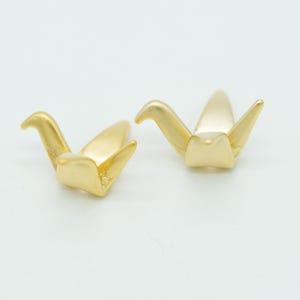 May include: Two small, matte gold-toned metal charms shaped like stylized origami cranes. Each charm features a pointed head, two outstretched wings, and a tapered tail section, with a smooth, uniform metallic finish across the entire surface.