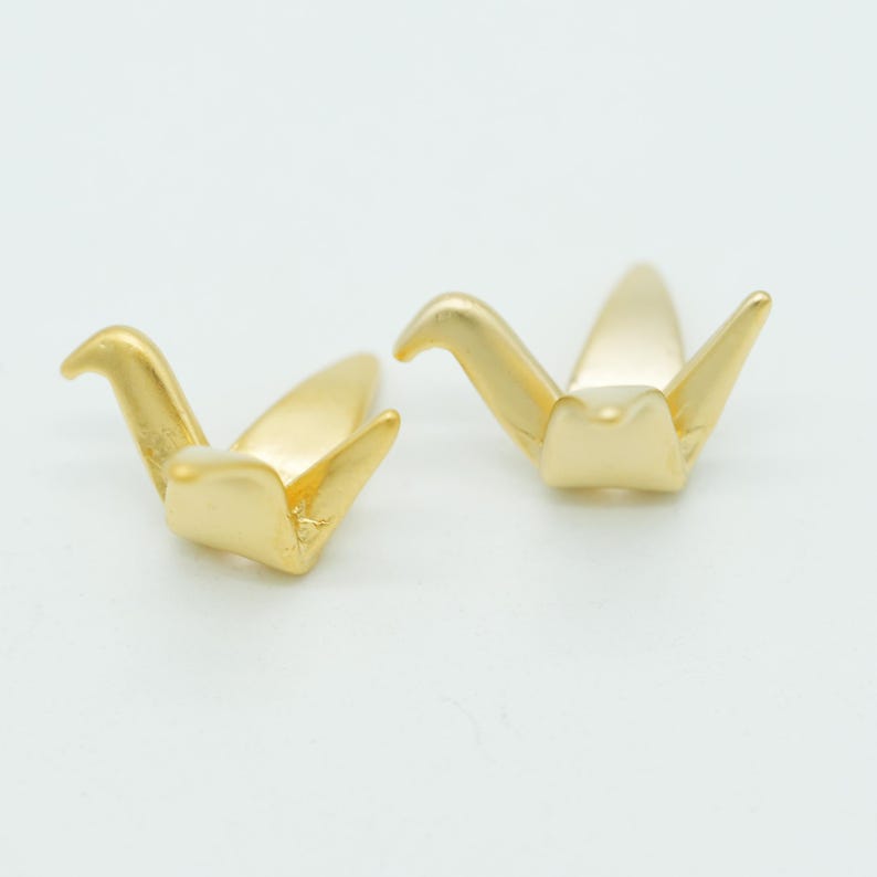 May include: Two small, matte gold-toned metal charms shaped like stylized origami cranes. Each charm features a pointed head, two outstretched wings, and a tapered tail section, with a smooth, uniform metallic finish across the entire surface.